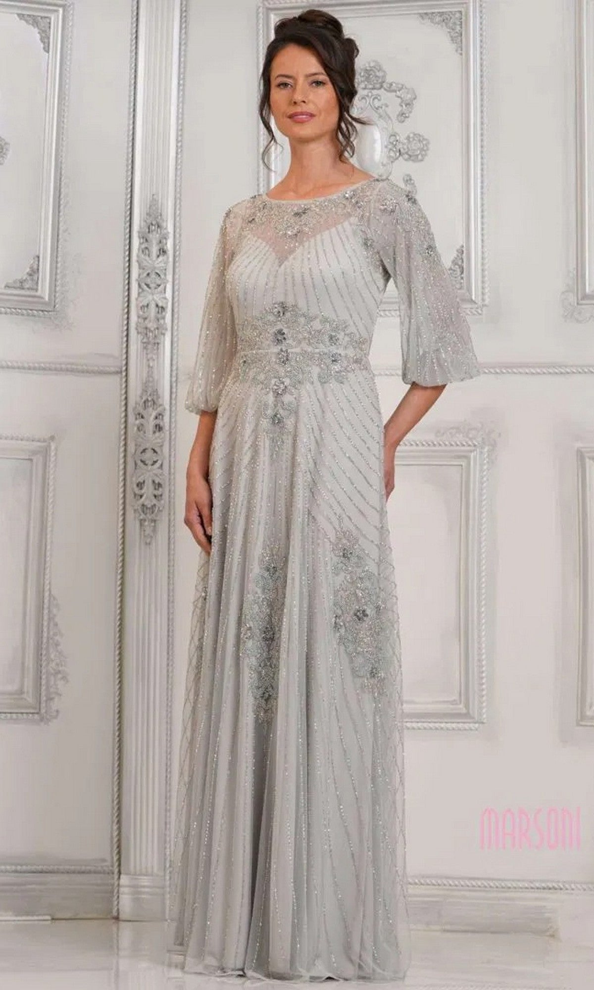 A woman in a silver Marsoni MV1323 long formal dress with sheer sleeves and intricate beading stands before an ornate white wall.